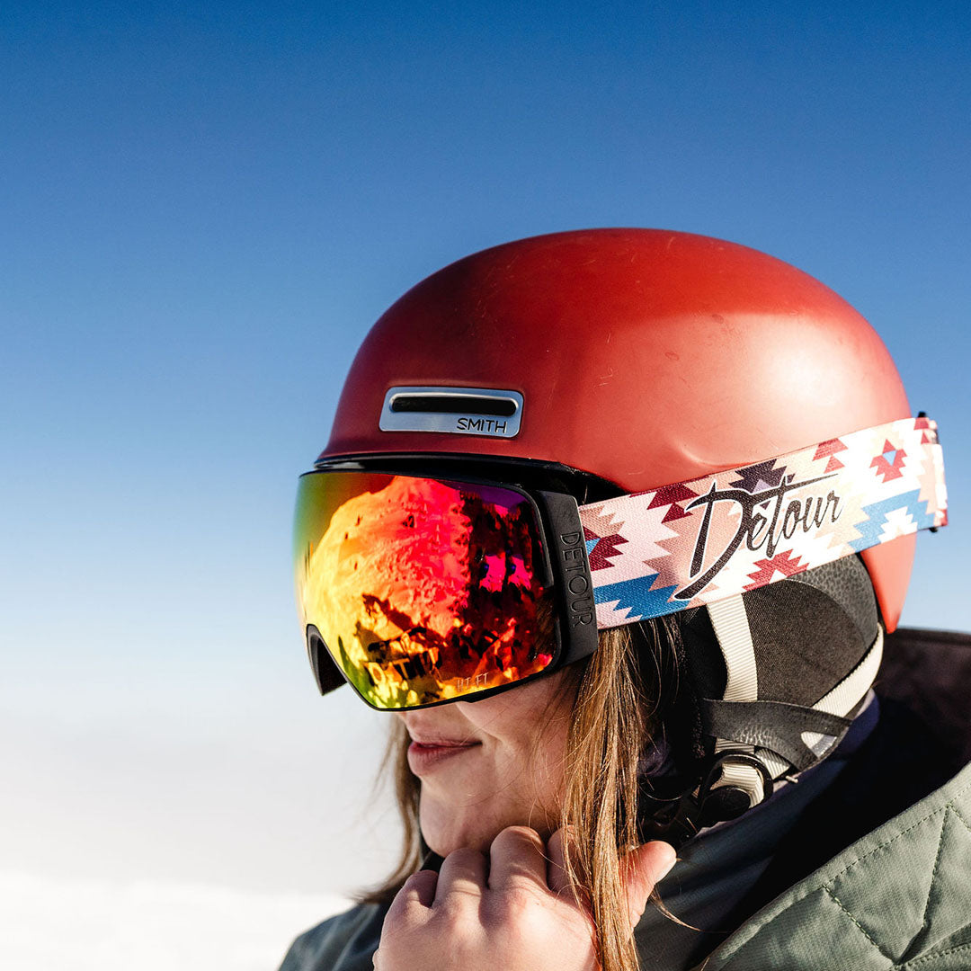 Snow goggles with reflective blue lens half-buried in snow, 'Detour' logo visible on strap.